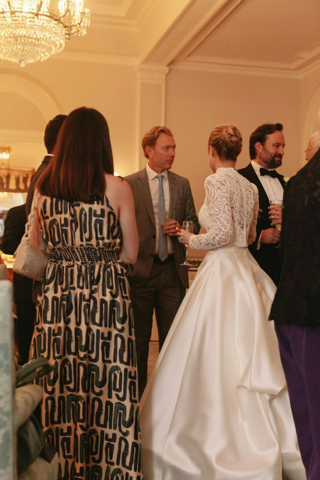 Group of people in formal attire at a wedding reception, including the bride in a white wedding gown and others holding drinks, engaging in conversation in an elegant, well-lit room with a chandelier.