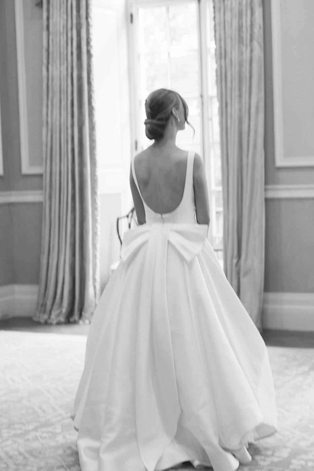 A woman in a wedding dress standing indoors near a large window with curtains.