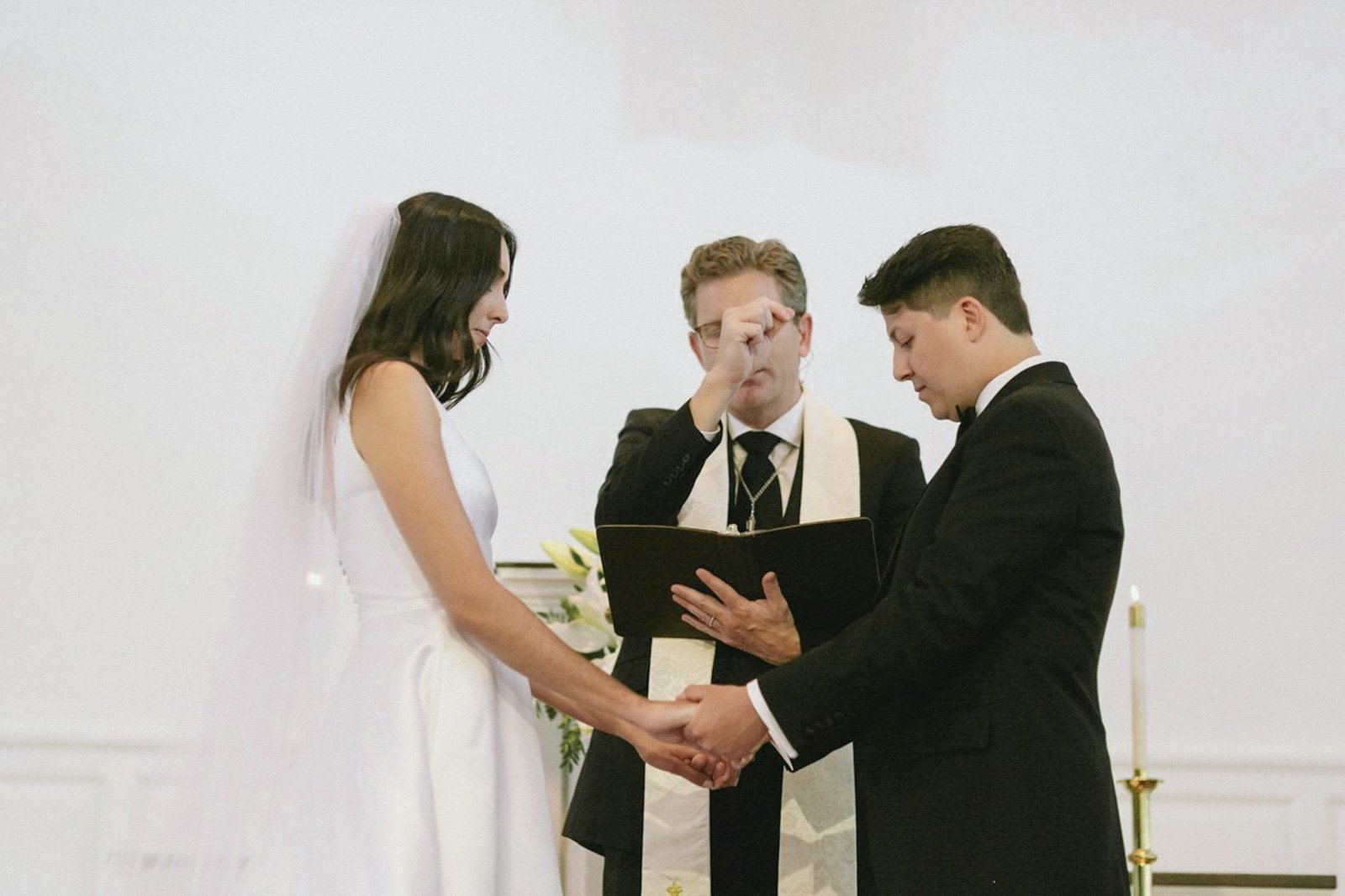 A bride and groom holding hands during a wedding ceremony, with an officiant reading from a book.