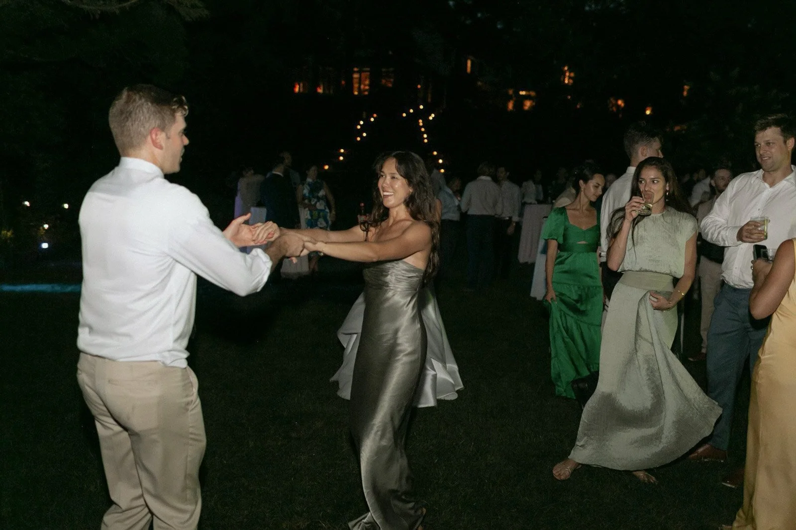 People dancing and socializing outdoors at night, with festive lights in the background.