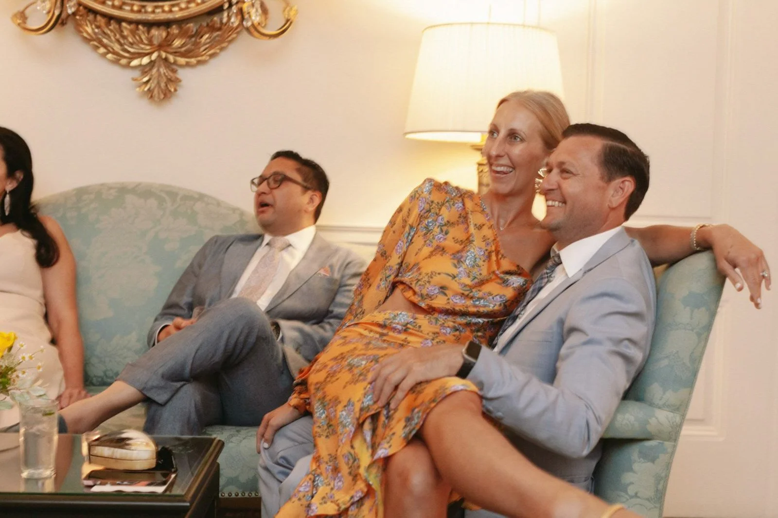 People sitting on a light blue sofa, smiling and laughing, in a well-lit room with a lamp and botanical wall decor at The Colony Club New York.
