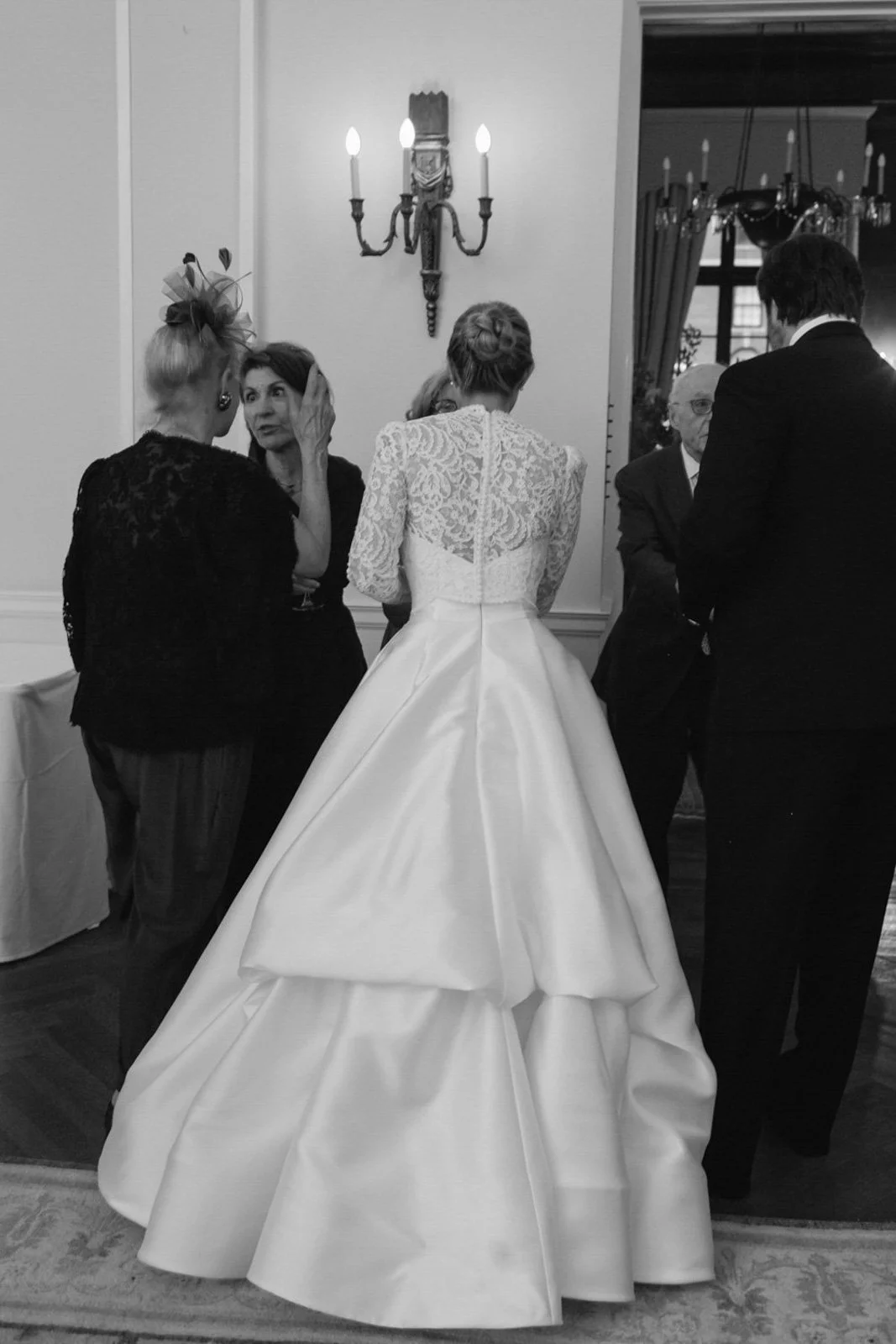 A woman in a luxurious white wedding dress with lace on the upper part and a voluminous skirt, seen from the back. She is in a room with ornate wall sconces and chandeliers, engaged in conversation with several other well-dressed women and an older m