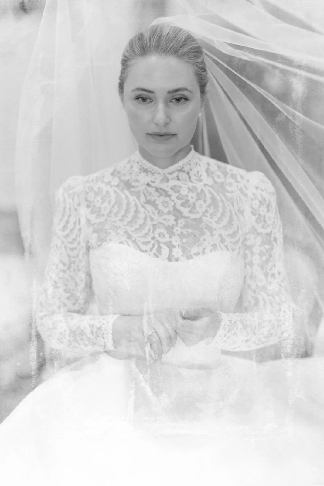A woman with blonde hair in an updo wearing a high-collared lace dress with long sleeves, standing in front of light-colored fabric or curtains.