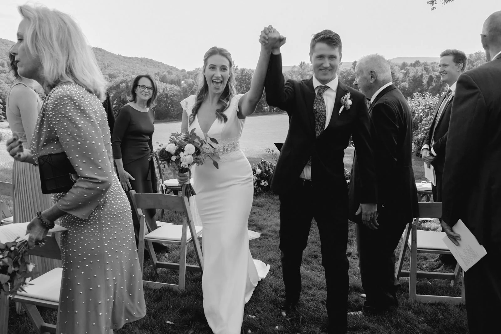 A bride and groom holding hands and smiling at an outdoor wedding ceremony, surrounded by attendees.