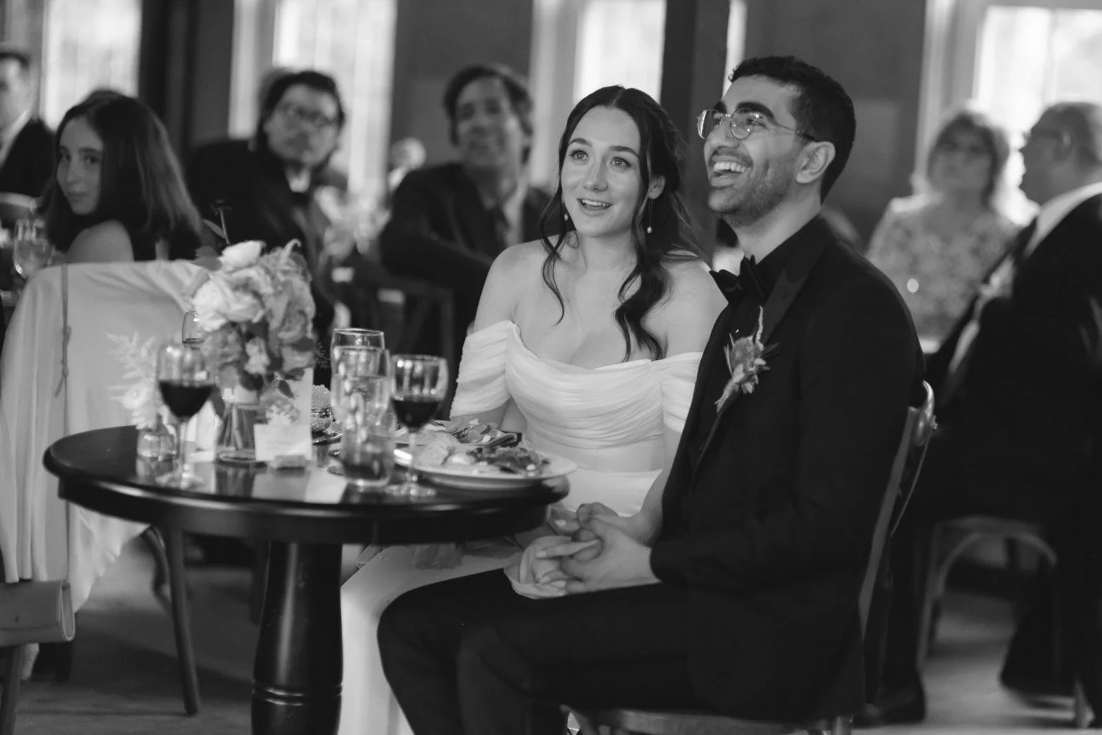 A bride and groom sitting at a table during a wedding reception, smiling and holding hands. The bride is wearing an off-the-shoulder white gown, and the groom is in a tuxedo. Other guests are seated nearby, and the scene is in black and white.