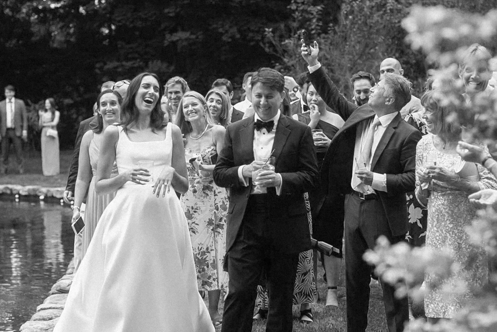 A black and white photo of a group of people in formal attire at an outdoor event, smiling and holding drinks, near a pond with trees in the background.