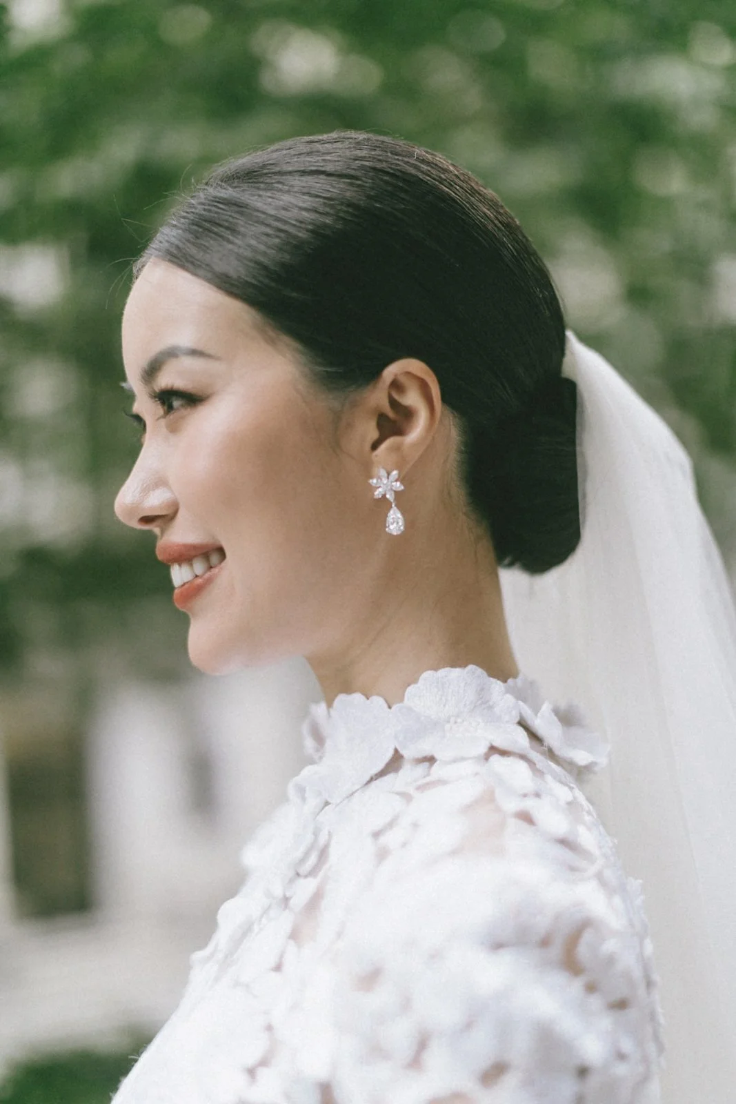 A woman with dark hair styled in a neat bun, wearing bridal jewelry and a white lace wedding dress, smiling outdoors with blurred green background.