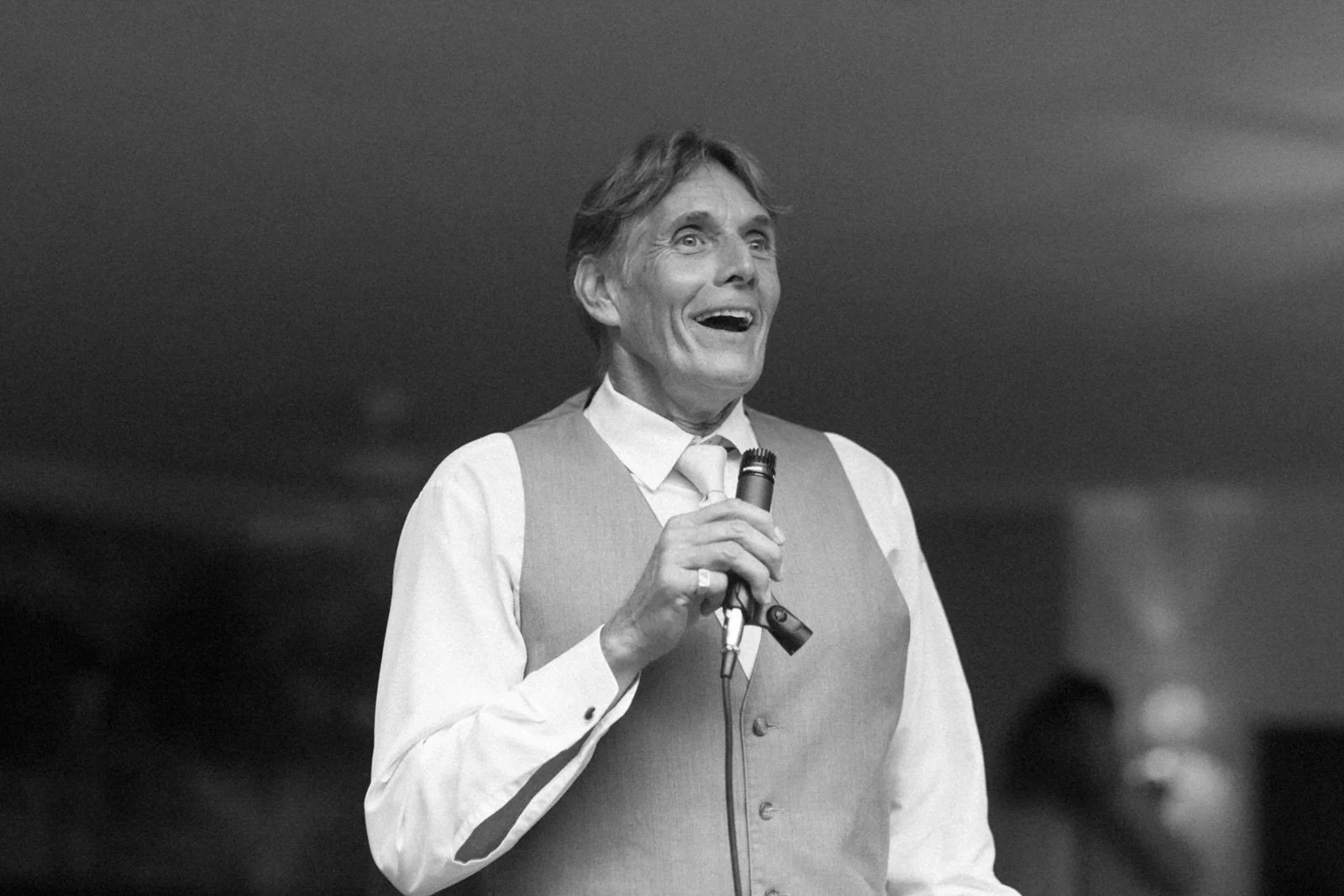 Black and white photo of a man in a vest and tie holding a microphone, smiling with his mouth open, in front of a dark background.
