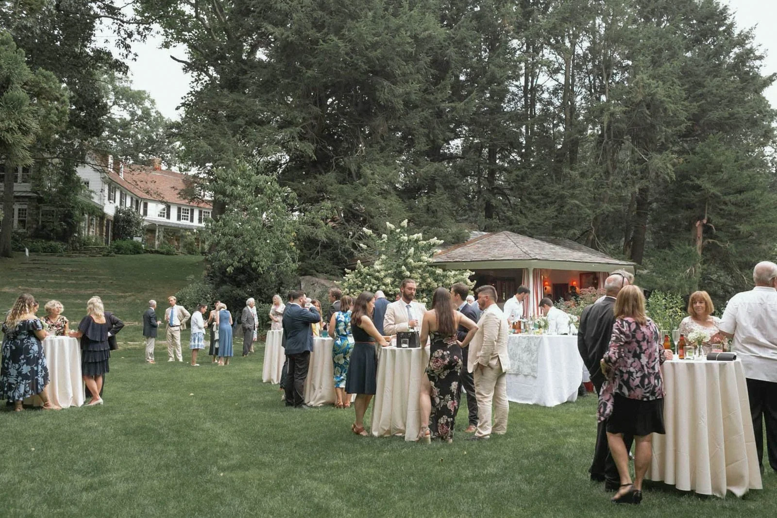 A group of people at an outdoor wedding reception or party on a grassy lawn with tall trees and large houses in the background. Attendees are standing around cocktail tables, socializing, and enjoying drinks.