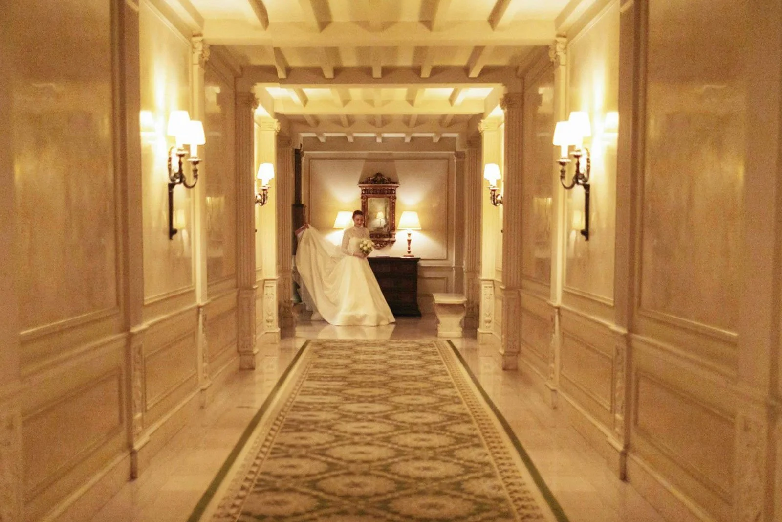 A bride in a white wedding dress holding a bouquet, standing in a luxurious, warmly lit hallway with ornate wall sconces, wood-paneled walls, and a patterned carpet, near a person adjusting her train.