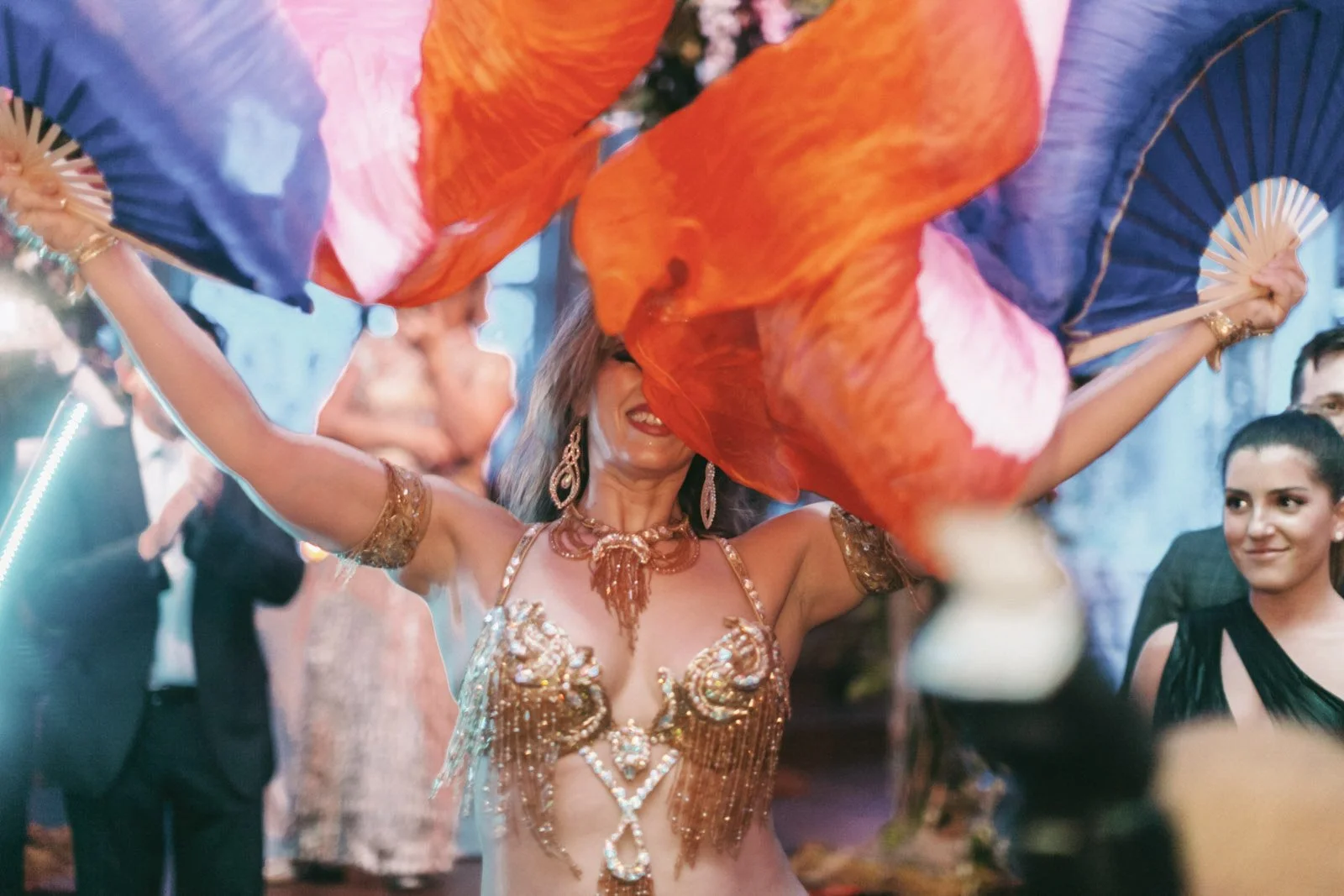 Woman in a sparkling gold costume dancing with colorful fans at a party.