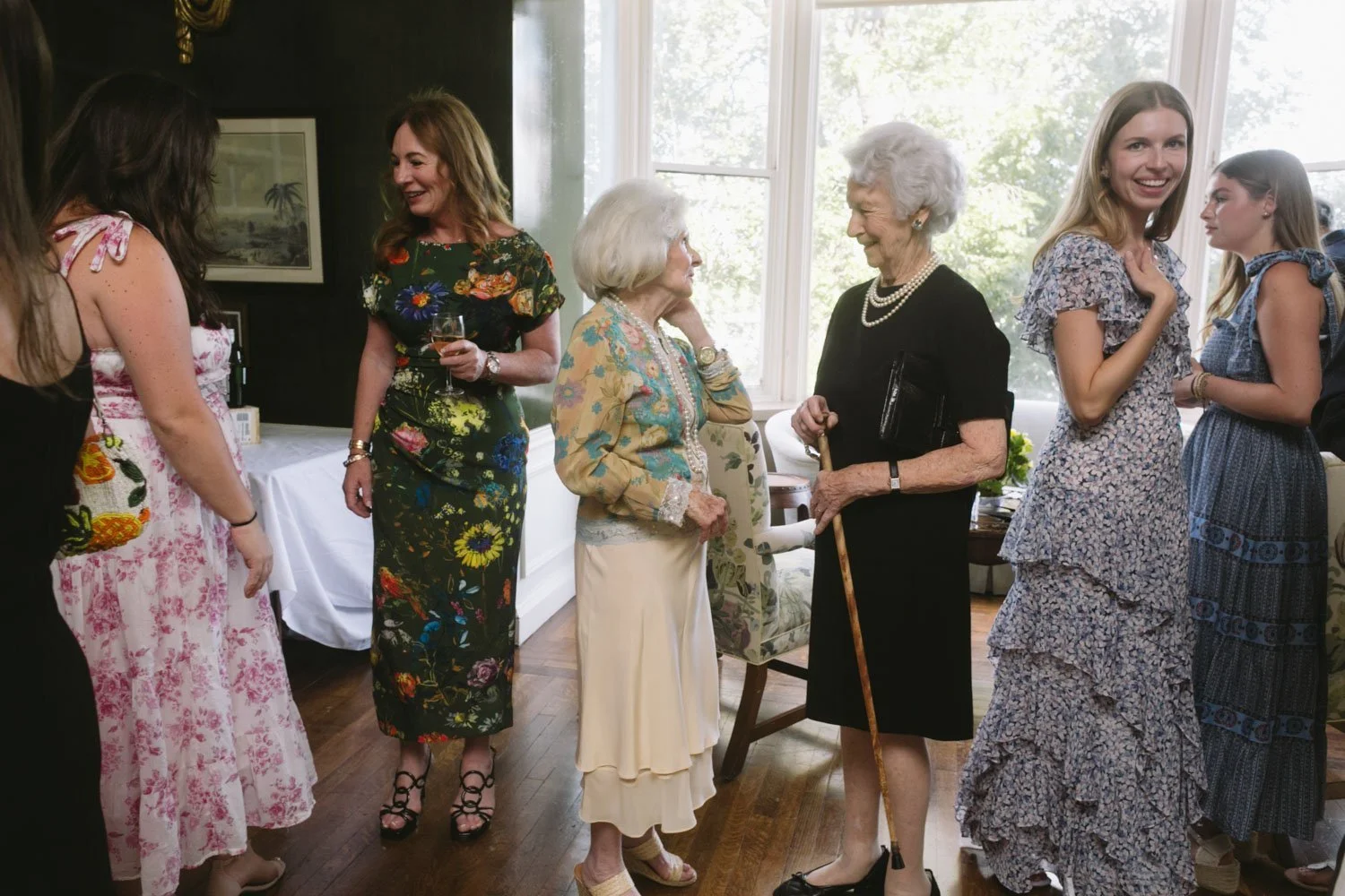 Group of women of different ages socializing indoors, some holding wine glasses, with sunlight coming through large windows behind them.