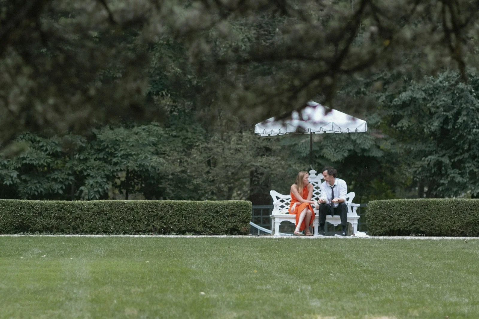 A man and woman sitting on a white bench under a white patio umbrella in a garden, engaged in conversation, with grass and hedges in the foreground.