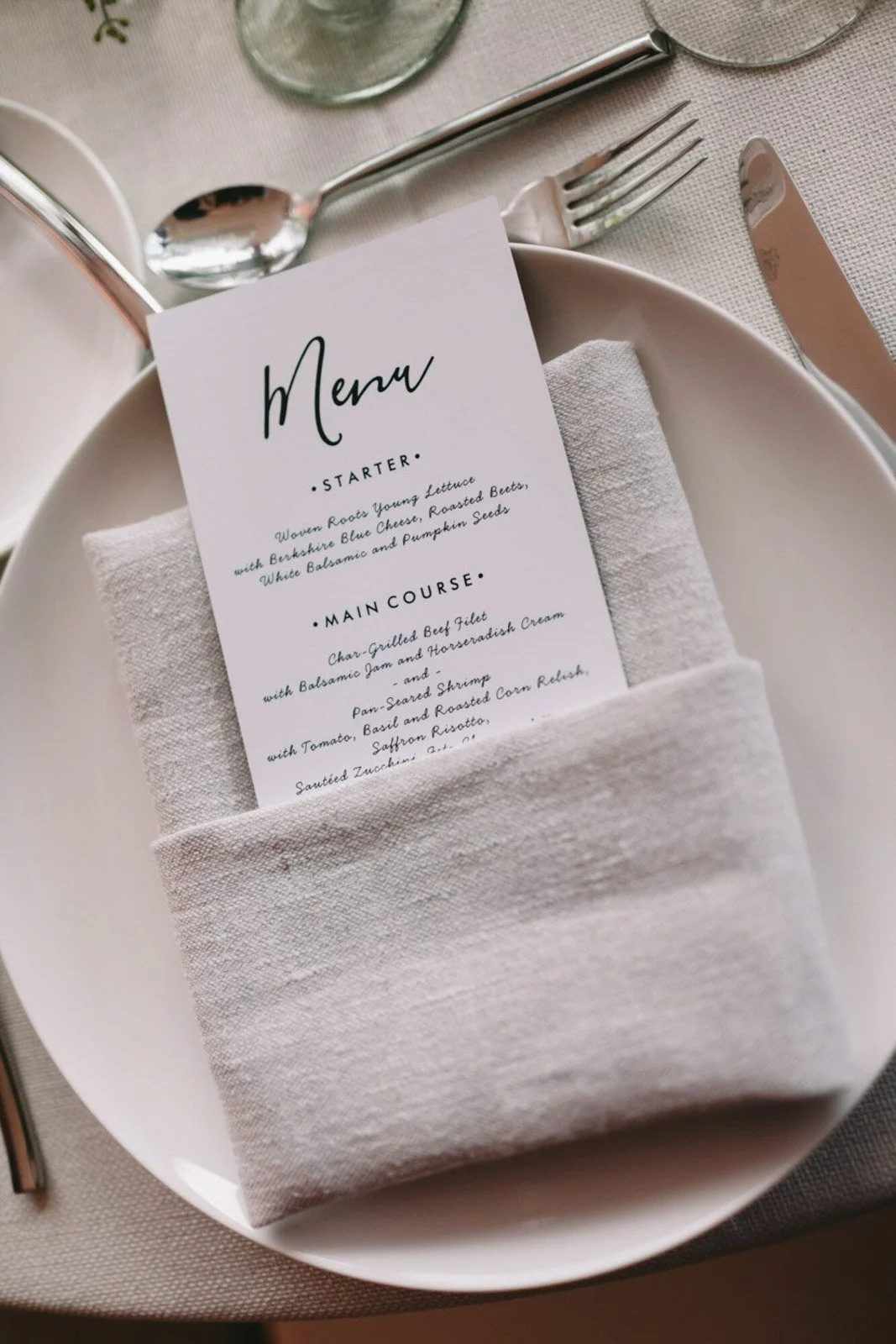 A printed menu resting in a folded cloth napkin on a white plate, with a fork, knife, and spoon arranged nearby on a table with a beige tablecloth.