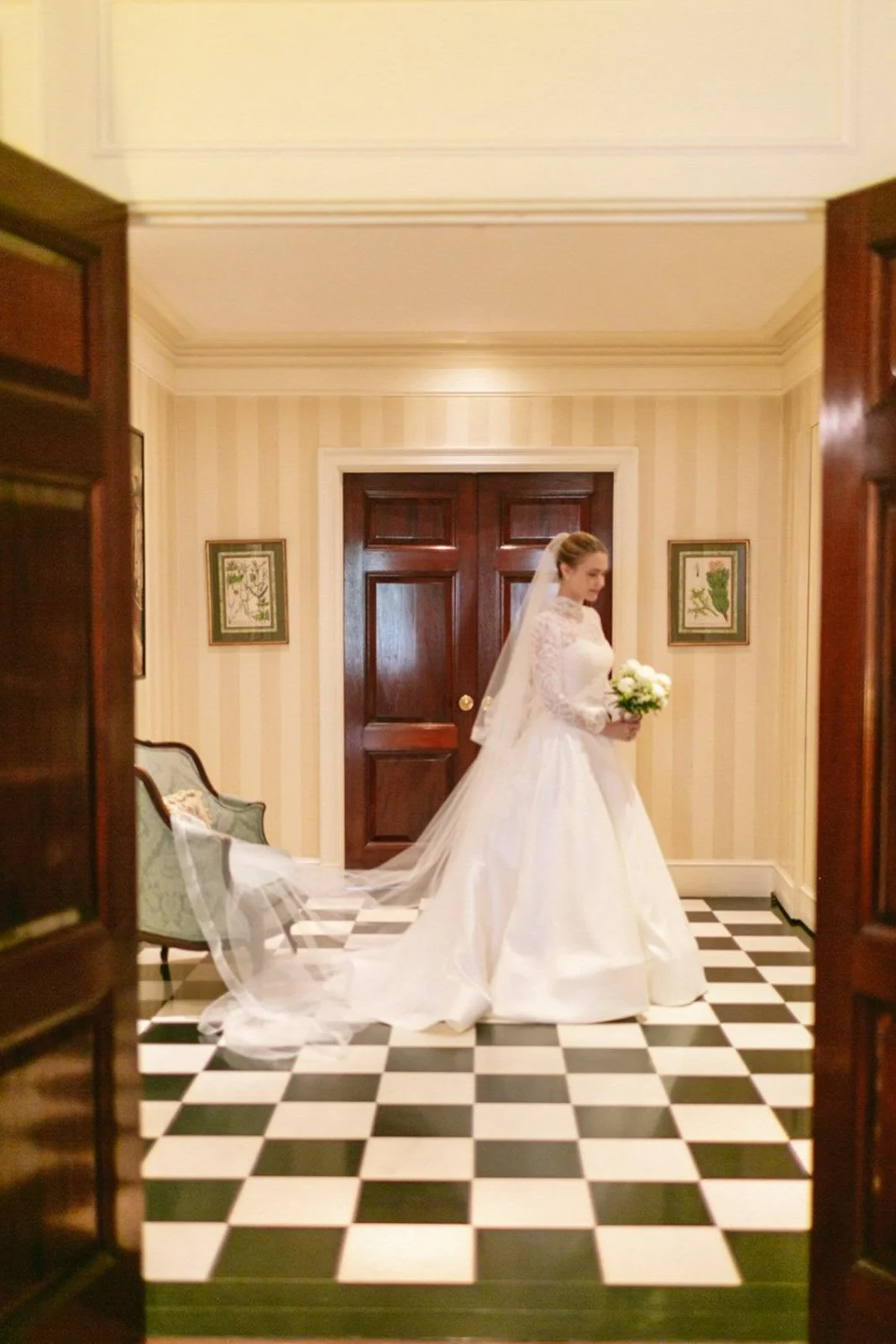 A bride in a white wedding gown standing inside an elegantly decorated room, holding a bouquet of white flowers.