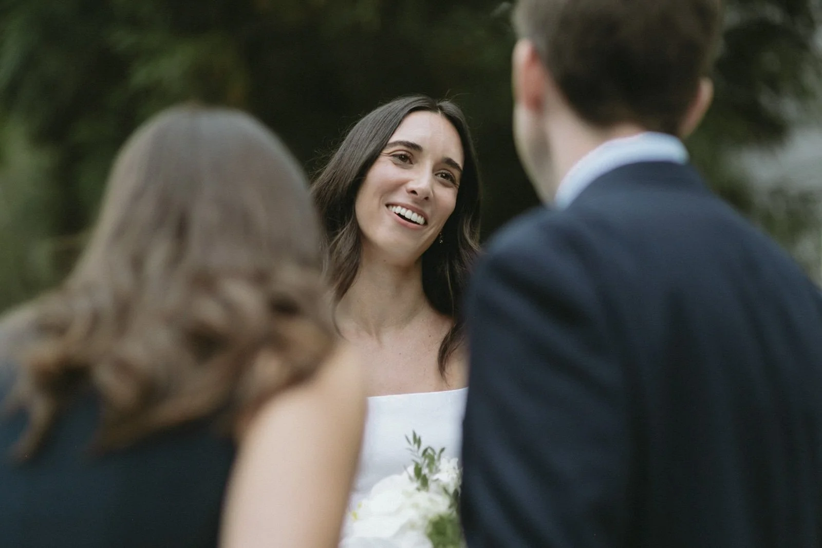 A woman with long dark hair, smiling, during a wedding ceremony with two other people, outdoors, with trees in the background.