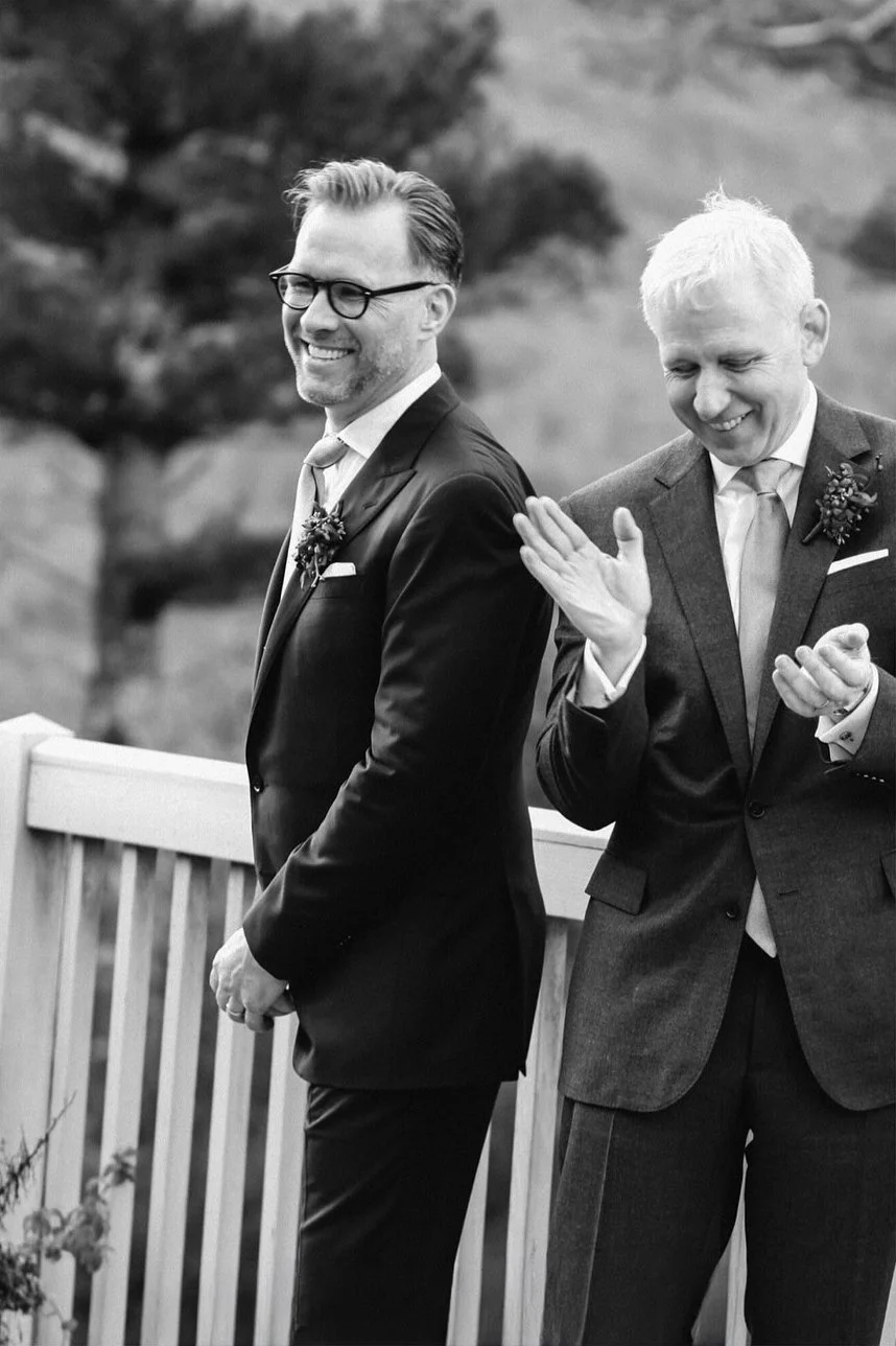 Two men in suits smiling at a wedding ceremony, standing outdoors near a wooden fence with a tree in the background.