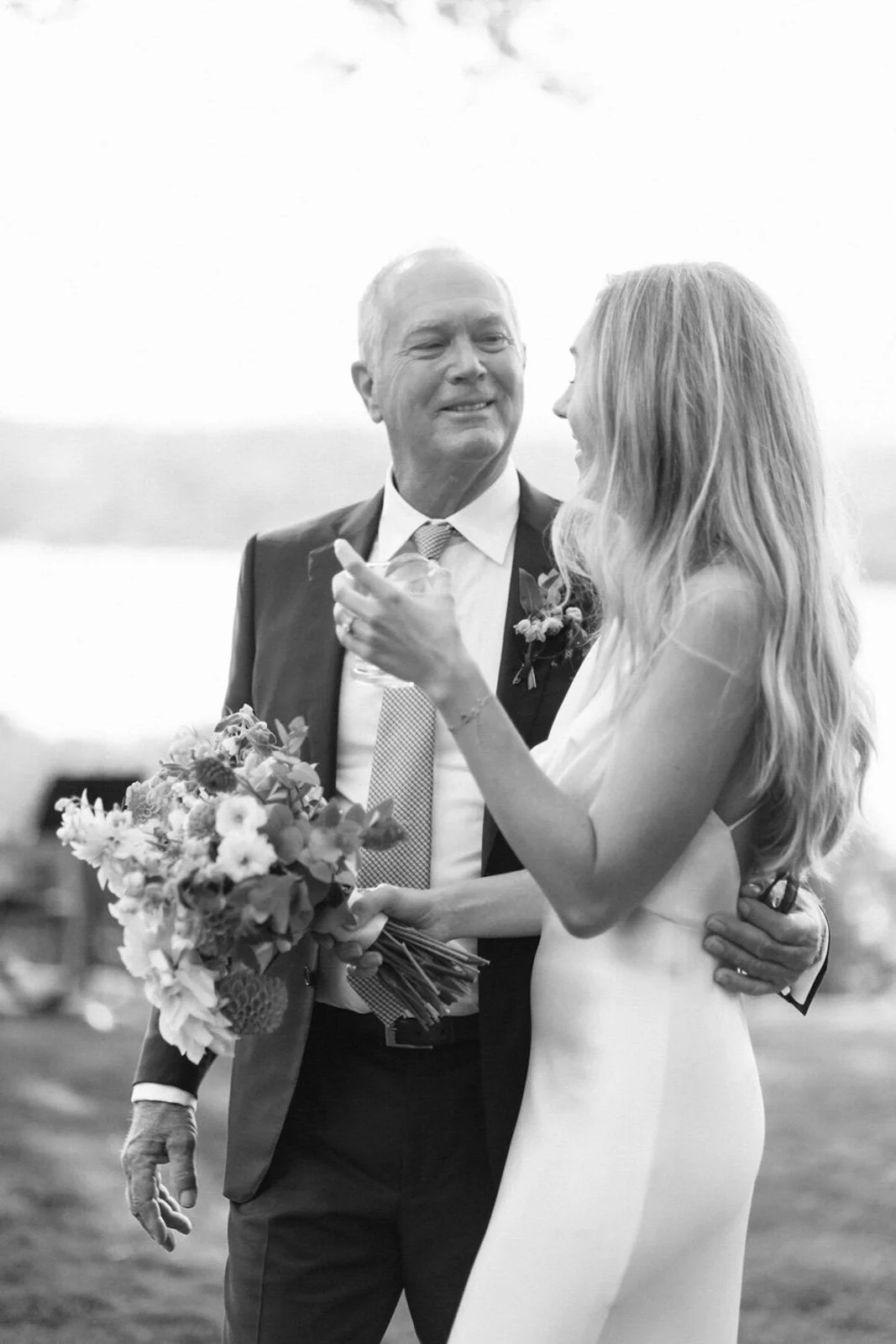 A black and white photo of a bride and an older man, likely her father, smiling at each other during a wedding. They are outside, with the bride holding a bouquet of flowers and wearing a wedding dress. The older man is in a suit with a boutonniere.