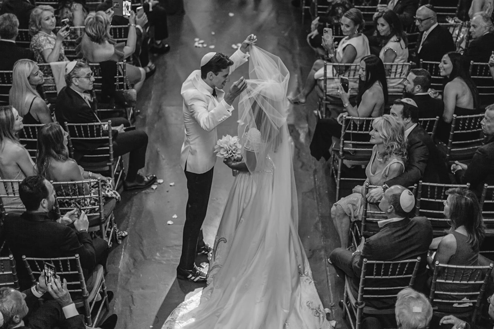A black-and-white photo of a wedding ceremony with a groom lifting a veil over the bride, surrounded by seated guests.
