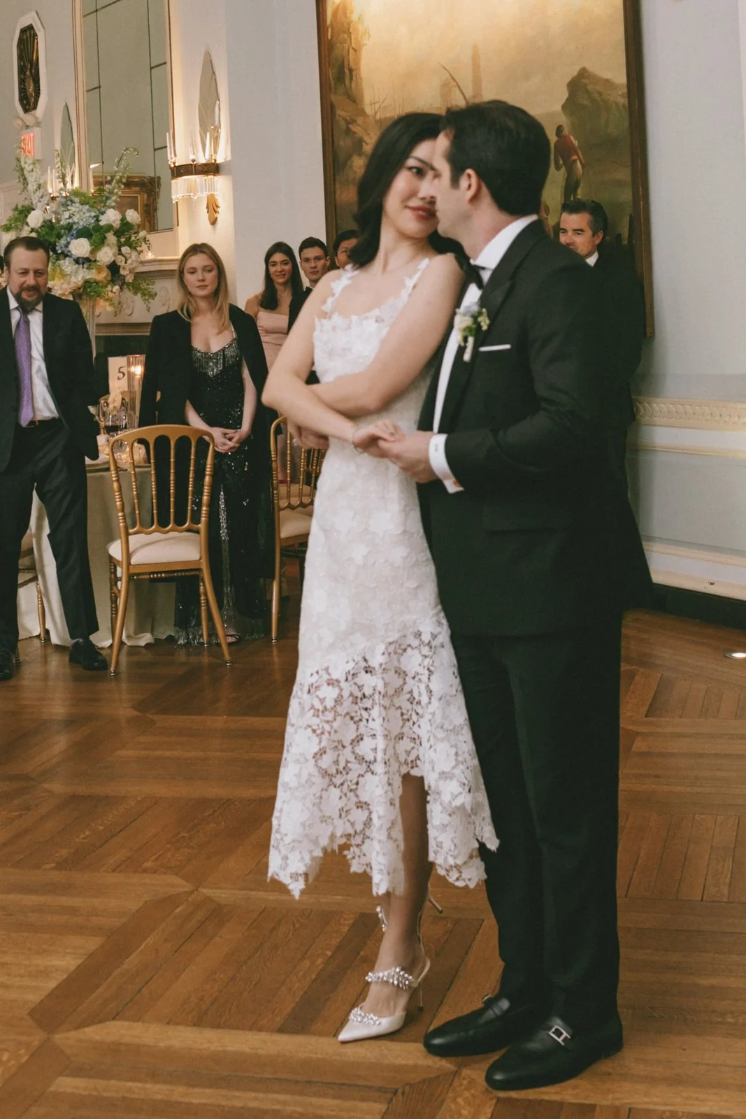 A bride and groom are dancing at their wedding reception, surrounded by guests and floral arrangements.