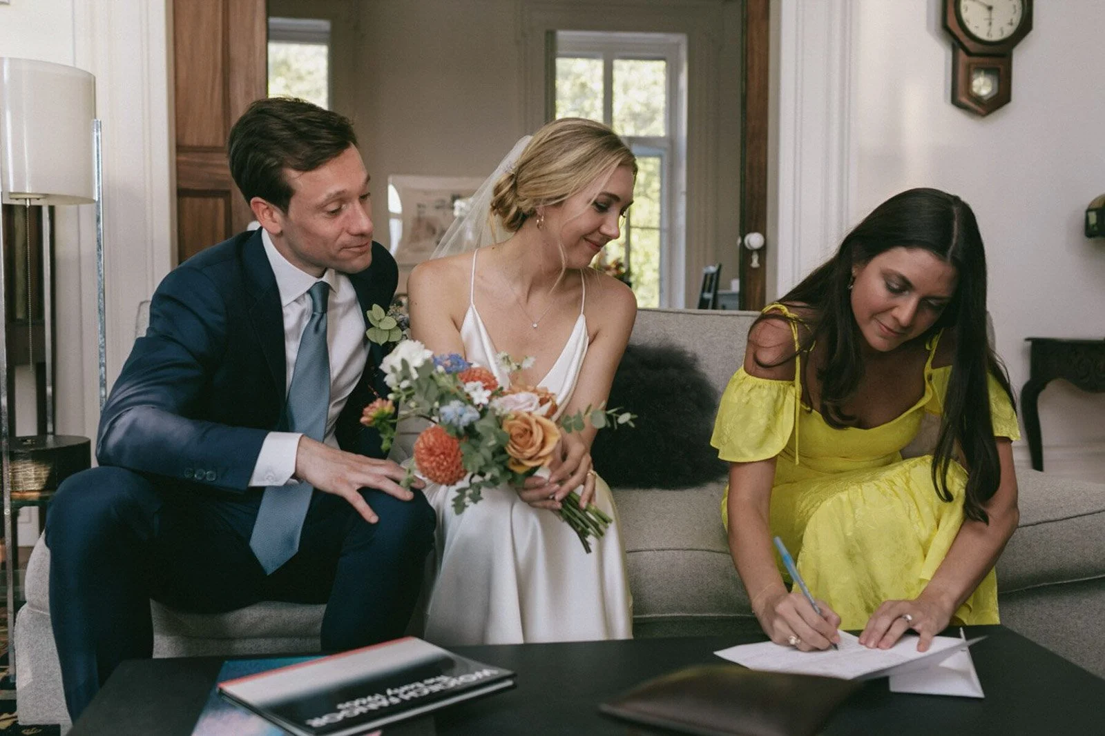 A bride in a white wedding dress holding a bouquet of flowers sitting on a sofa next to a man in a dark suit and light blue tie. A woman in a yellow dress is sitting on the other side, signing a document.