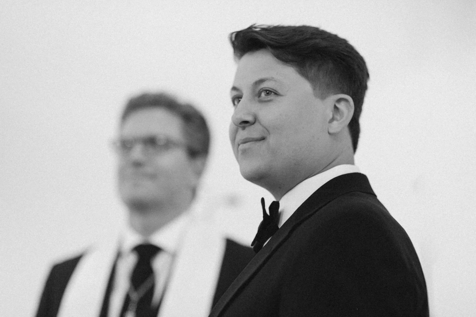 Black and white photo of two men in tuxedos, one in the foreground and one in the background, with the man in front smiling and the man in the background out of focus.