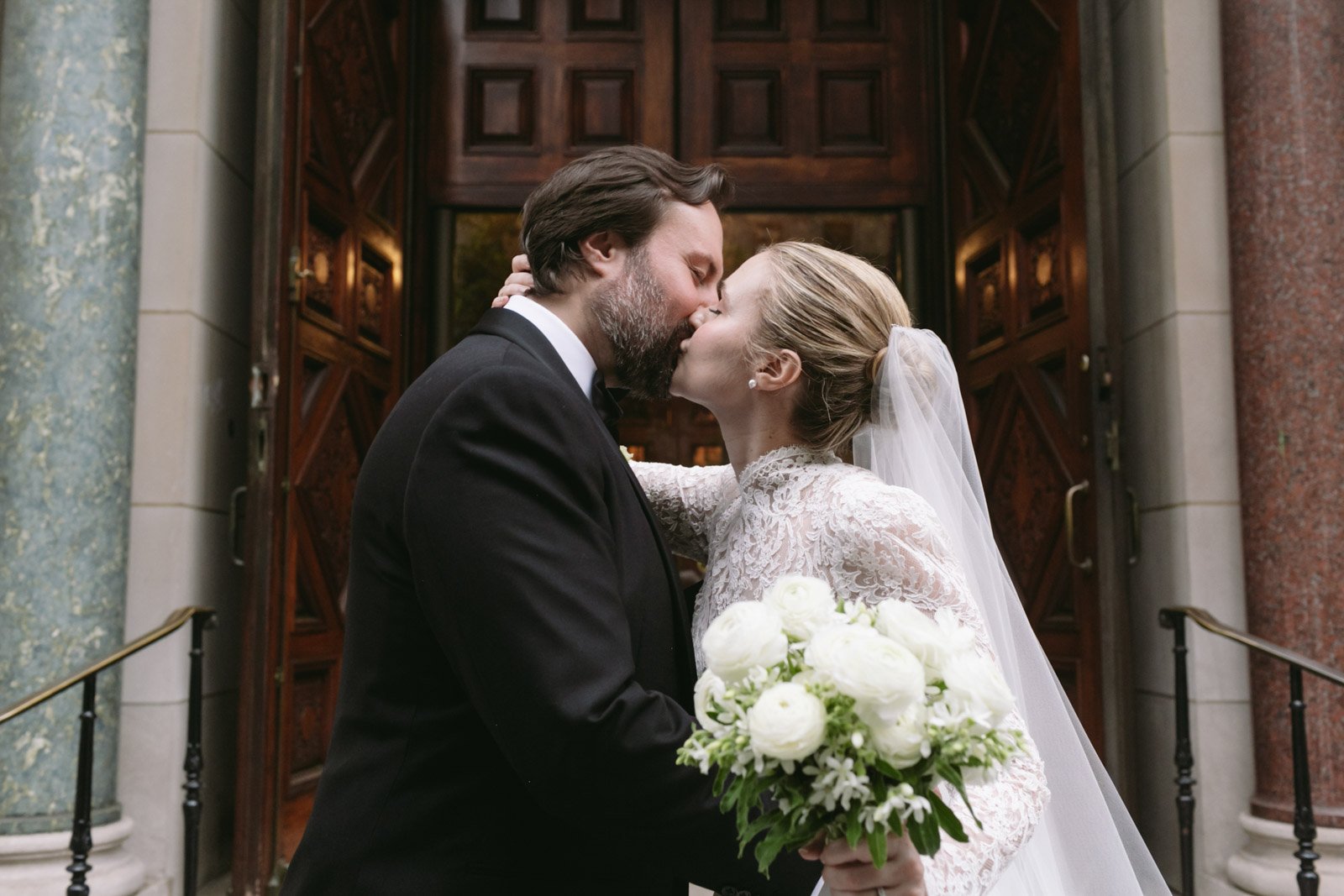 A bride and groom kissing in front of a church entrance, with the bride holding a bouquet of white flowers.