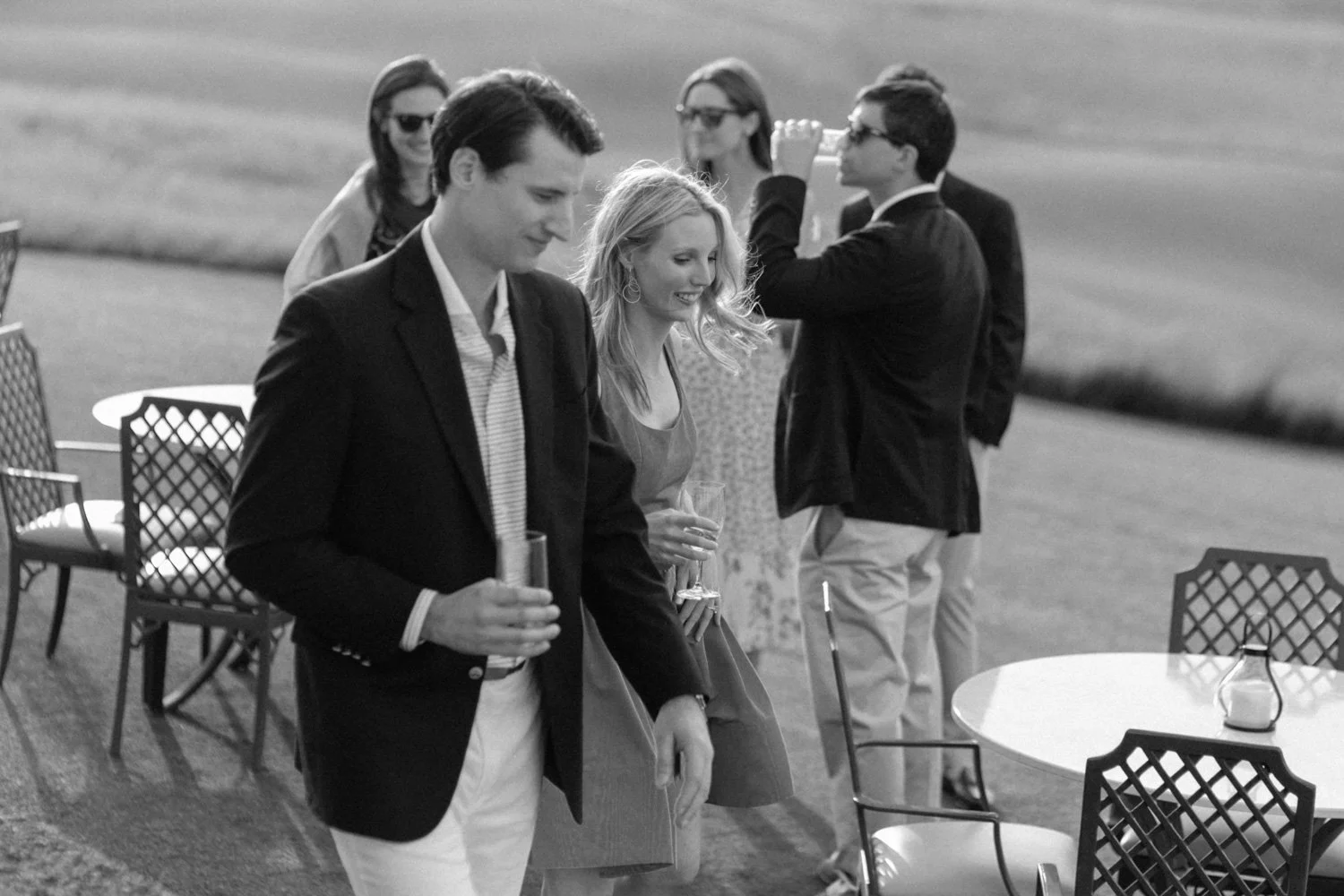 Black and white photo of six young adults at an outdoor gathering, holding drinks and socializing. Some are wearing sunglasses, dressed in semi-formal attire, with tables and chairs around them.