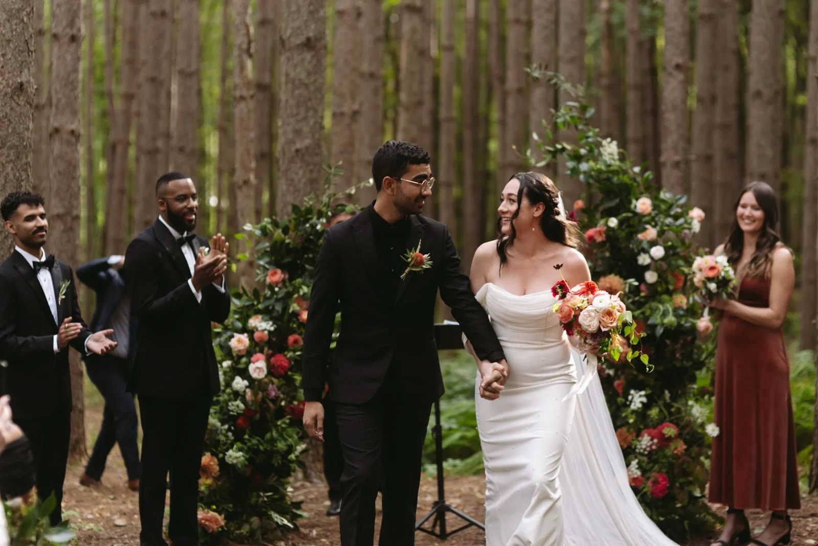 A bride and groom holding hands during their wedding ceremony in a forest, surrounded by friends and floral arrangements.