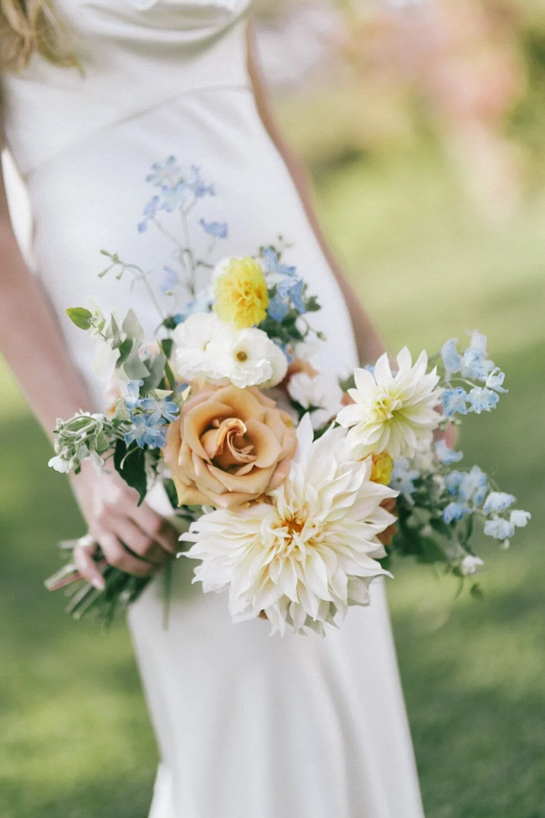 A bride holding a bouquet of white, peach, and blue flowers, with a blurred outdoor background at A Private Estate in Germantown, NY.