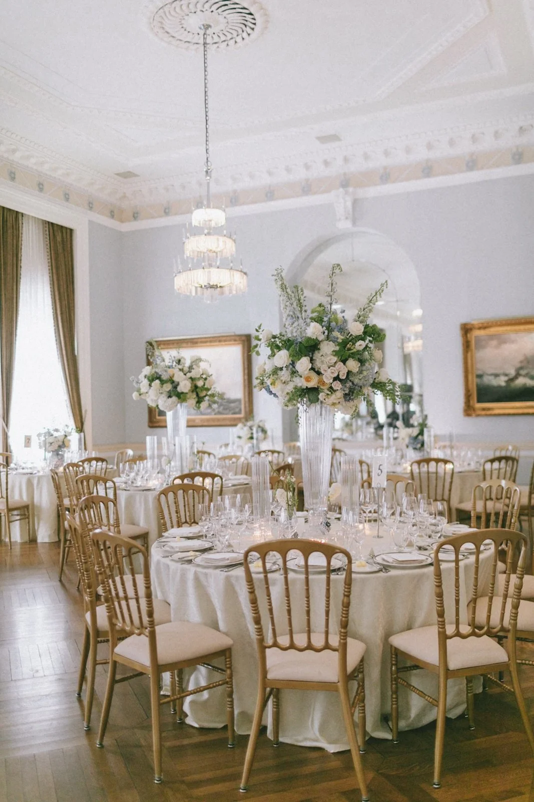 Elegant banquet room with round tables decorated with white tablecloths, fine china, glassware, and gold chairs. Each table has a tall vase with a large floral arrangement of white roses and greenery. The room features a high ceiling with a chandelie