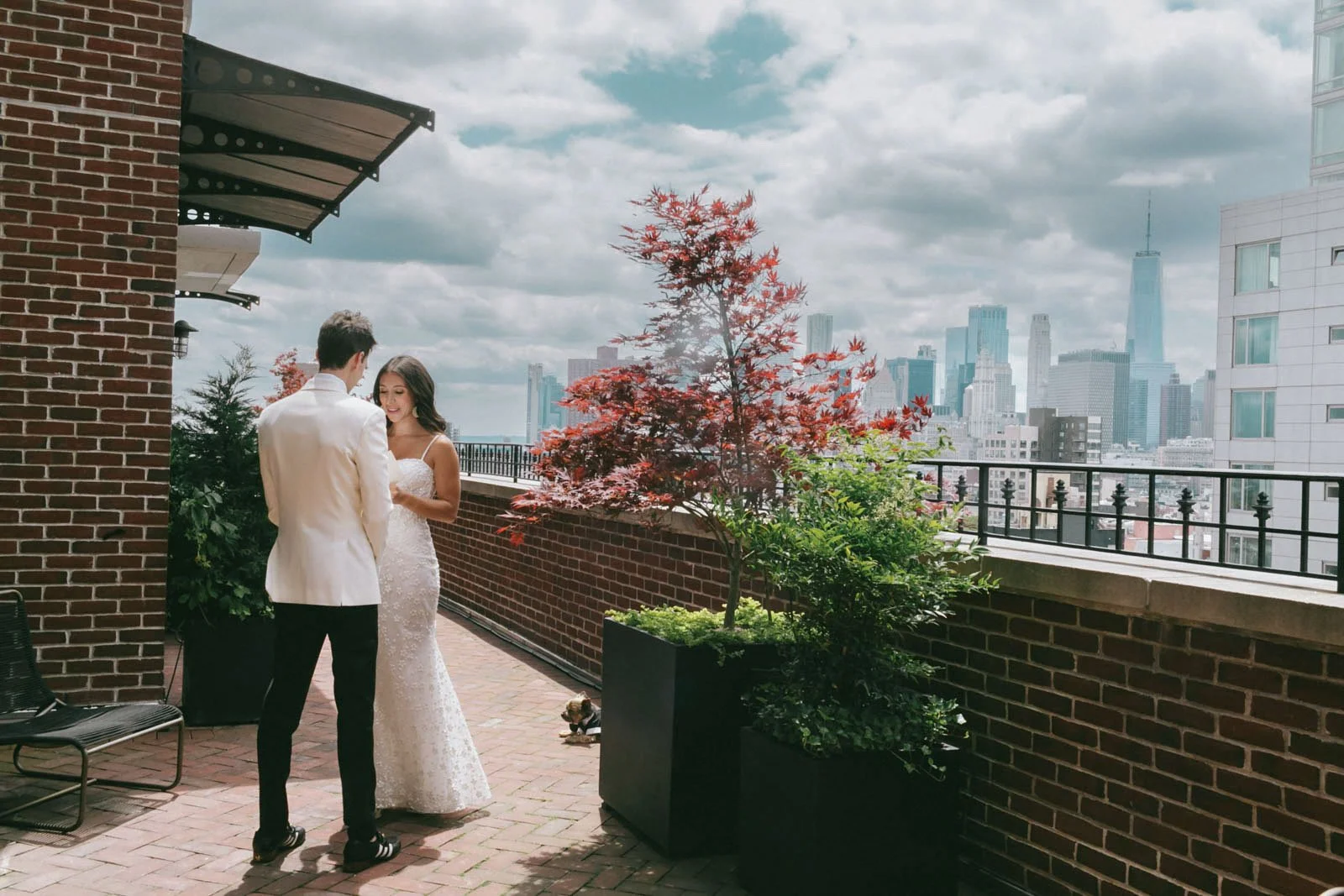 A couple dressed in wedding attire standing on a rooftop balcony with city skyline in the background. The woman wears a white lace wedding dress and the man a white suit jacket. There are potted plants and a black chair on the balcony, with cloudy sk