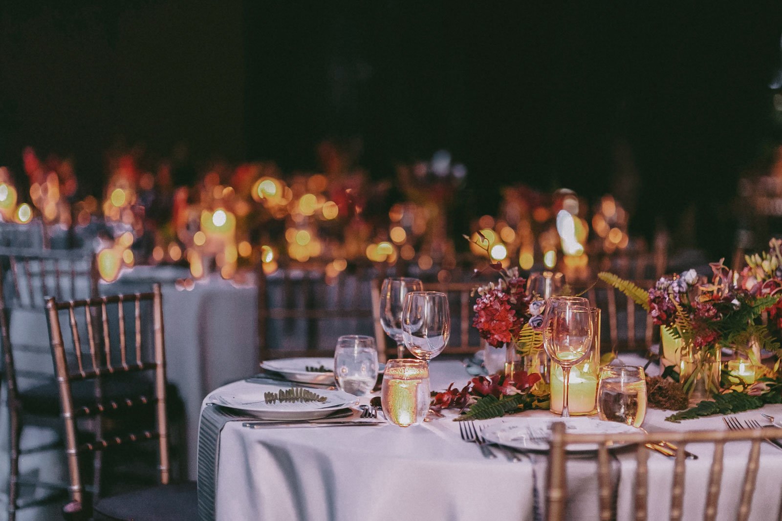 Elegant dining table set with glassware, floral arrangements, and candles at night, with blurred string lights in the background.