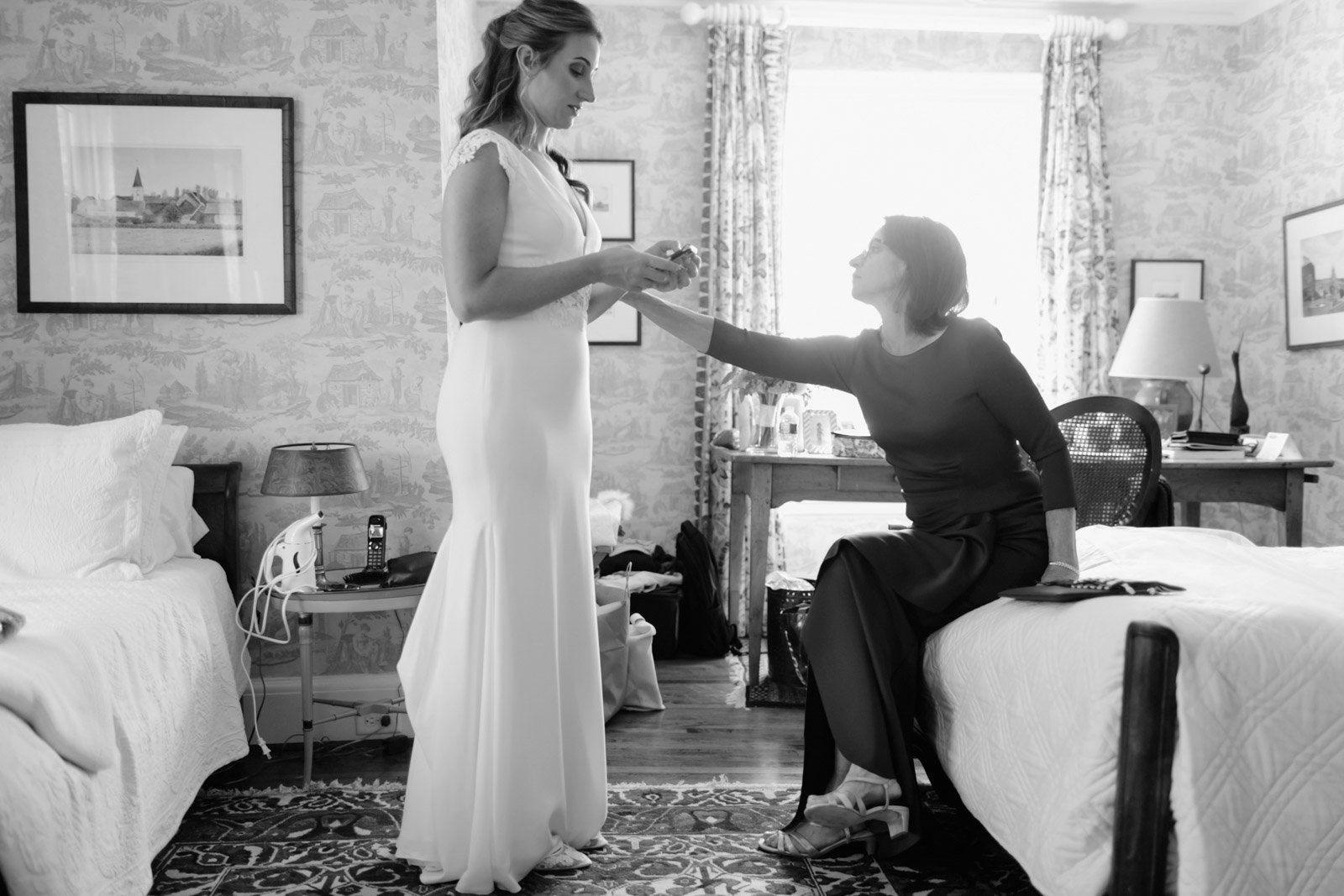 A bride in a white wedding gown stands in a hotel room while a woman, possibly her mother, sits on the bed reaching out to her hand. The room has patterned wallpaper, framed pictures on the wall, a desk with various items, and a bedside table with a 