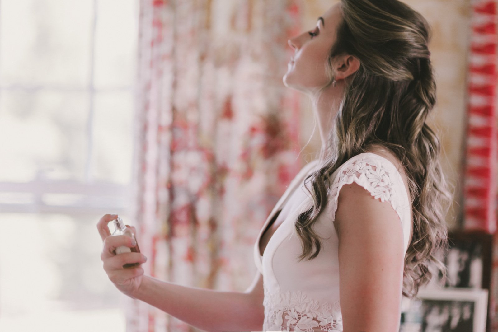 Woman spraying perfume in a softly lit room with floral curtains.