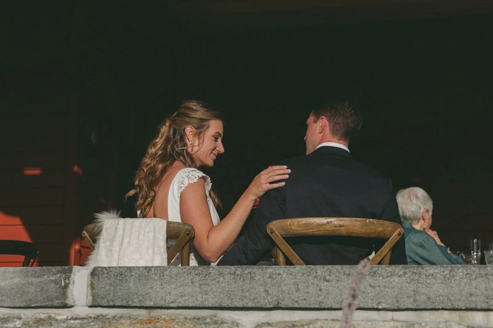 A bride and groom sitting closely together at a wedding reception, with an elderly woman in the background, under dim lighting.
