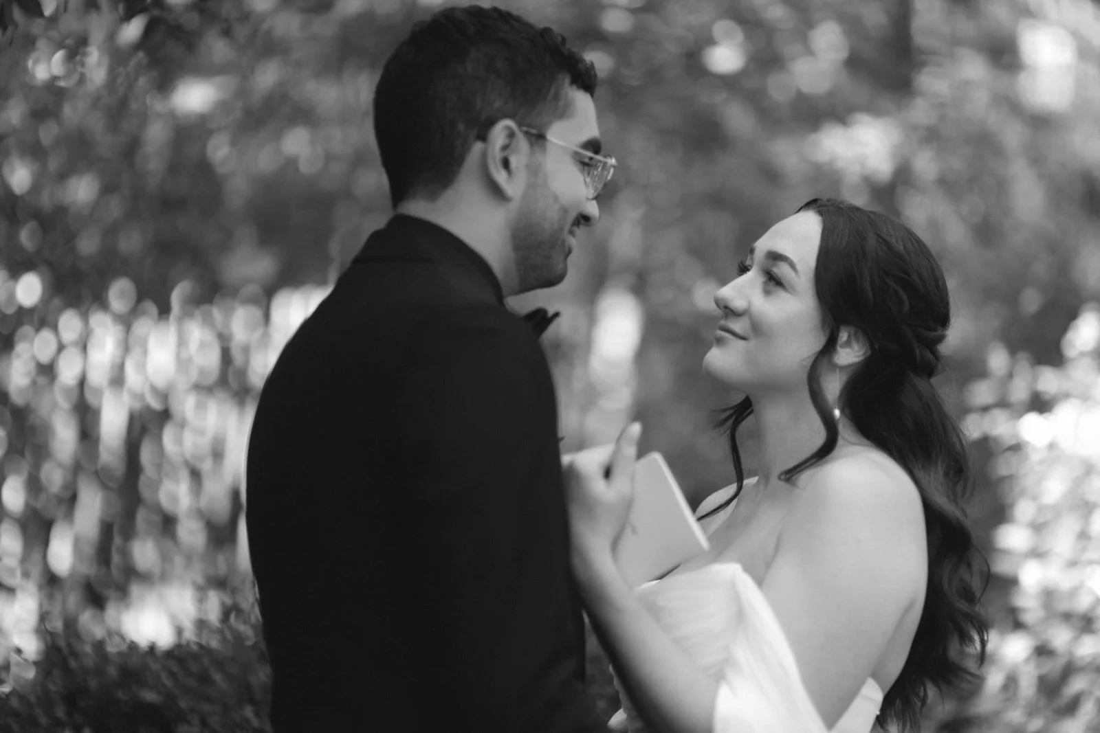 A couple dressed in wedding attire, standing outdoors, gazing at each other with smiles in a black-and-white photograph.