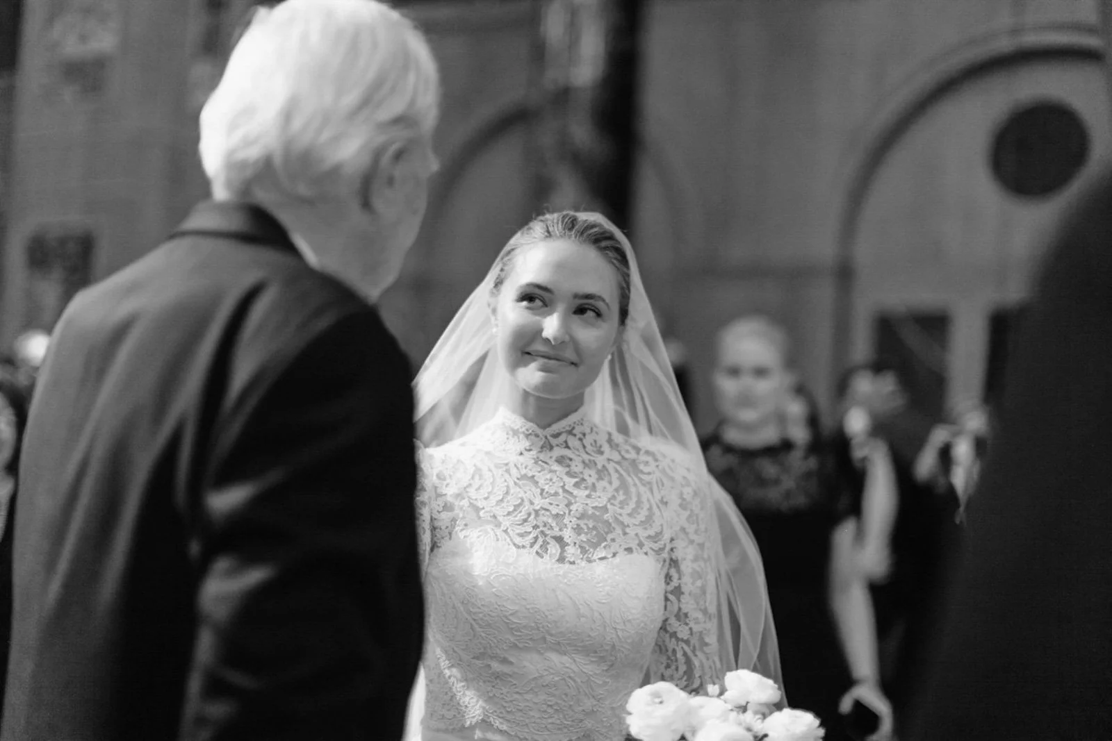 A bride in a lace wedding dress with a veil looking at an older man in a suit during a wedding ceremony in a church.