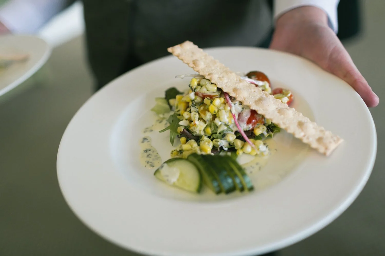 A plate with a salad containing corn, cherry tomatoes, and greens, topped with a crispy breadstick, with cucumber slices on the side.