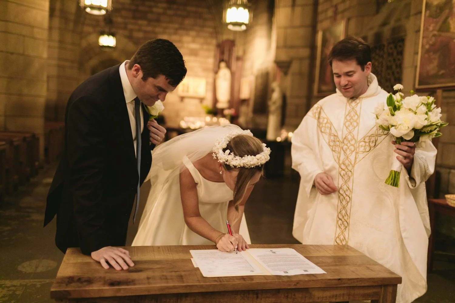 A bride signing a marriage document while two men, including a priest holding a bouquet, watch during a wedding ceremony in a church.