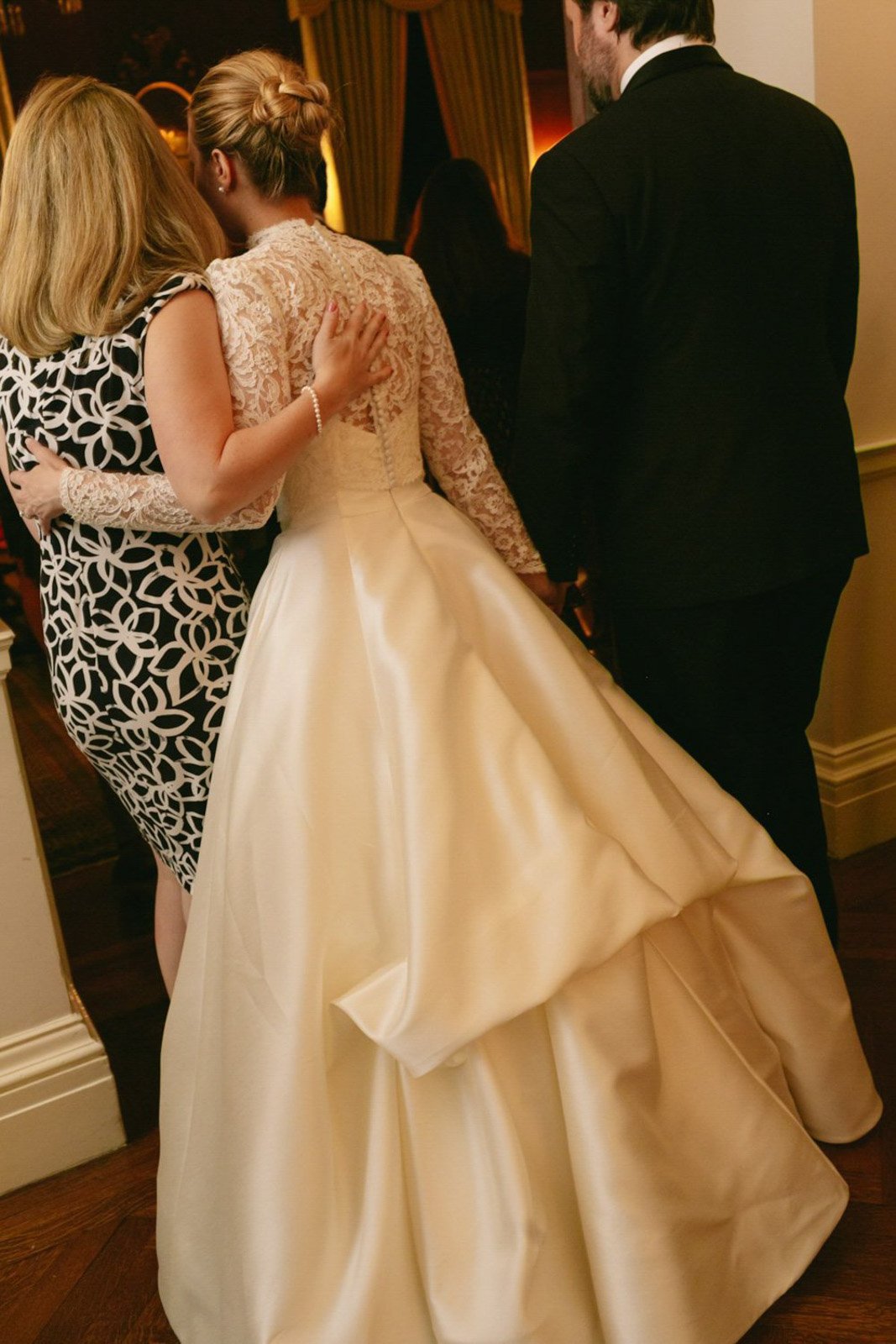 A bride in a white wedding gown with lace sleeves is hugging a woman in a black and white patterned dress. A man in a black suit stands nearby in a warmly lit room.