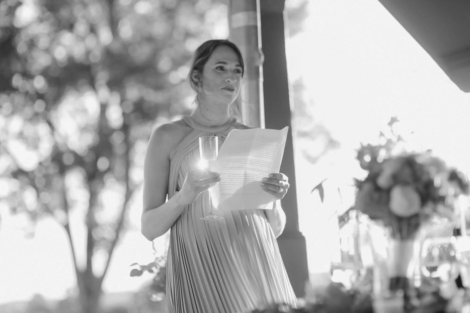 A woman in a sleeveless dress holding a glass of champagne and a piece of paper, possibly reading a speech, at a wedding reception or celebration, with trees in the background and sunlight shining through the window.