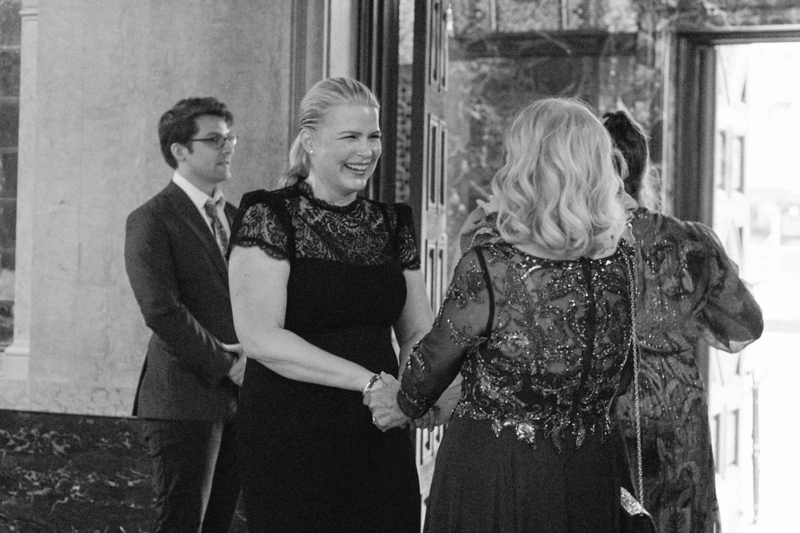 People at a semi-formal gathering, smiling and holding hands near a doorway in black and white.