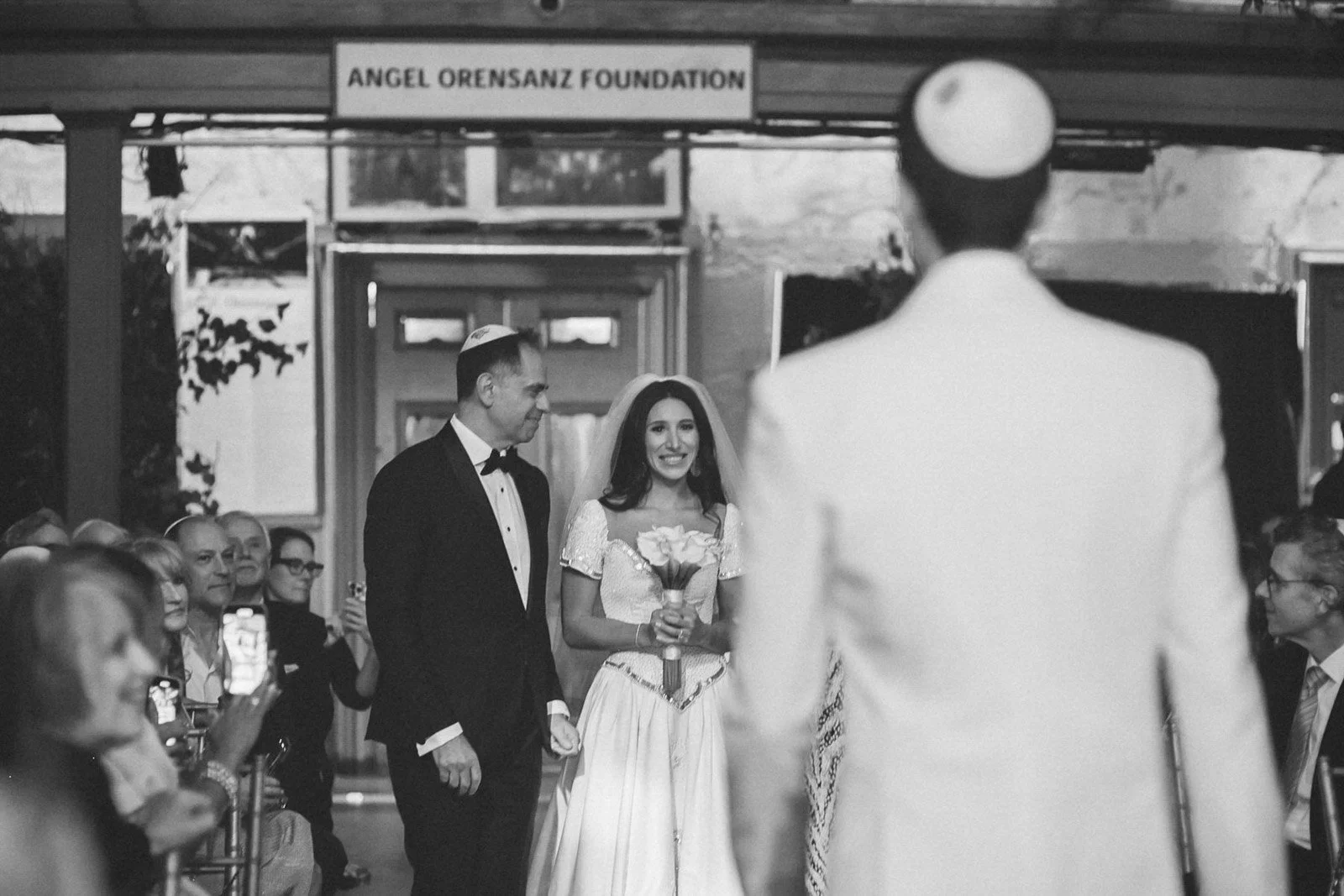 A black and white photo of a wedding ceremony, with a bride holding a bouquet, standing next to a man in a tuxedo. They are facing a man in a white suit and hat. Guests are seated on both sides, some taking photos. A sign reading 'Angel Orensanz Foun