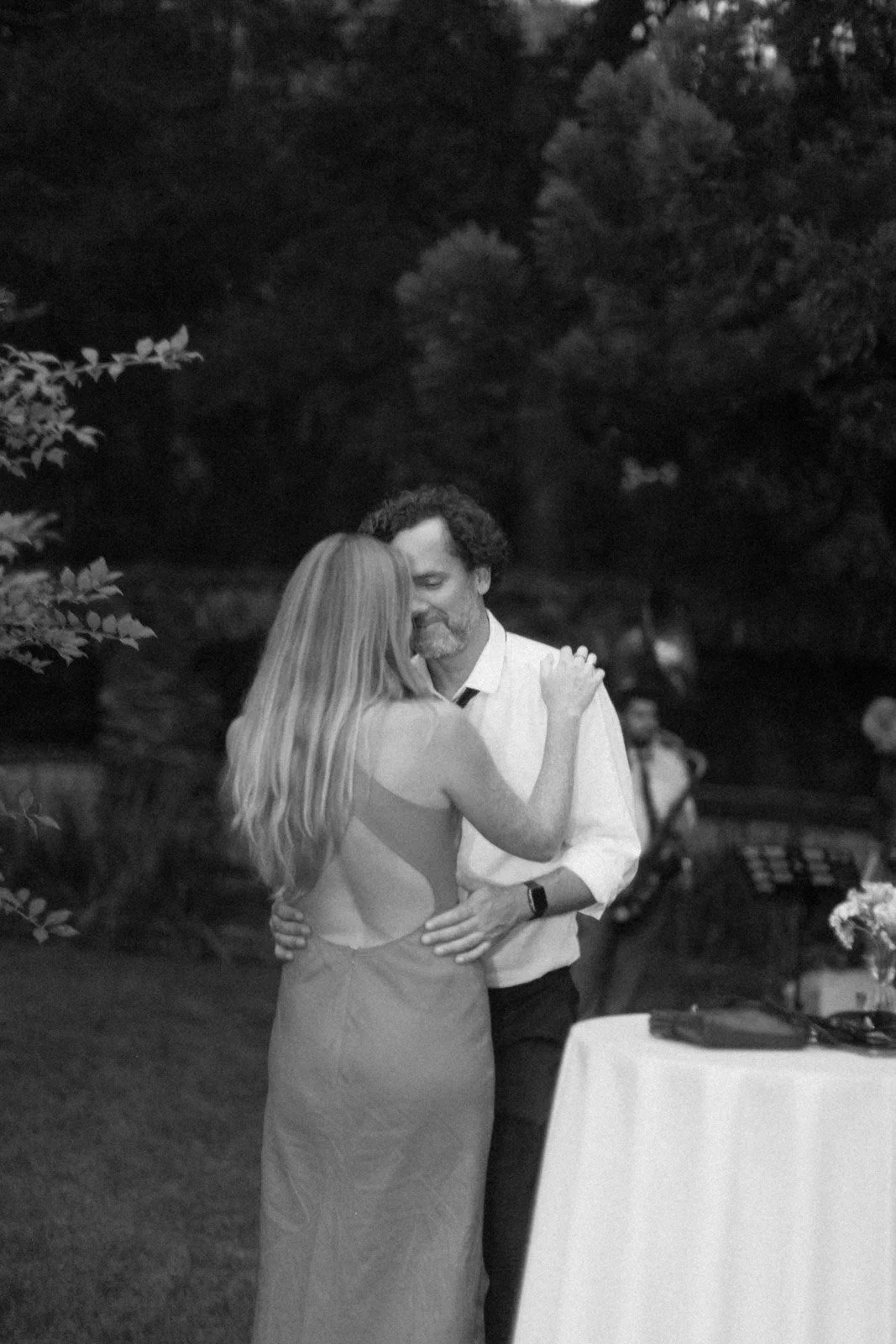 A black-and-white photo of a couple dancing closely in an outdoor setting at night, with trees and blurred people in the background.