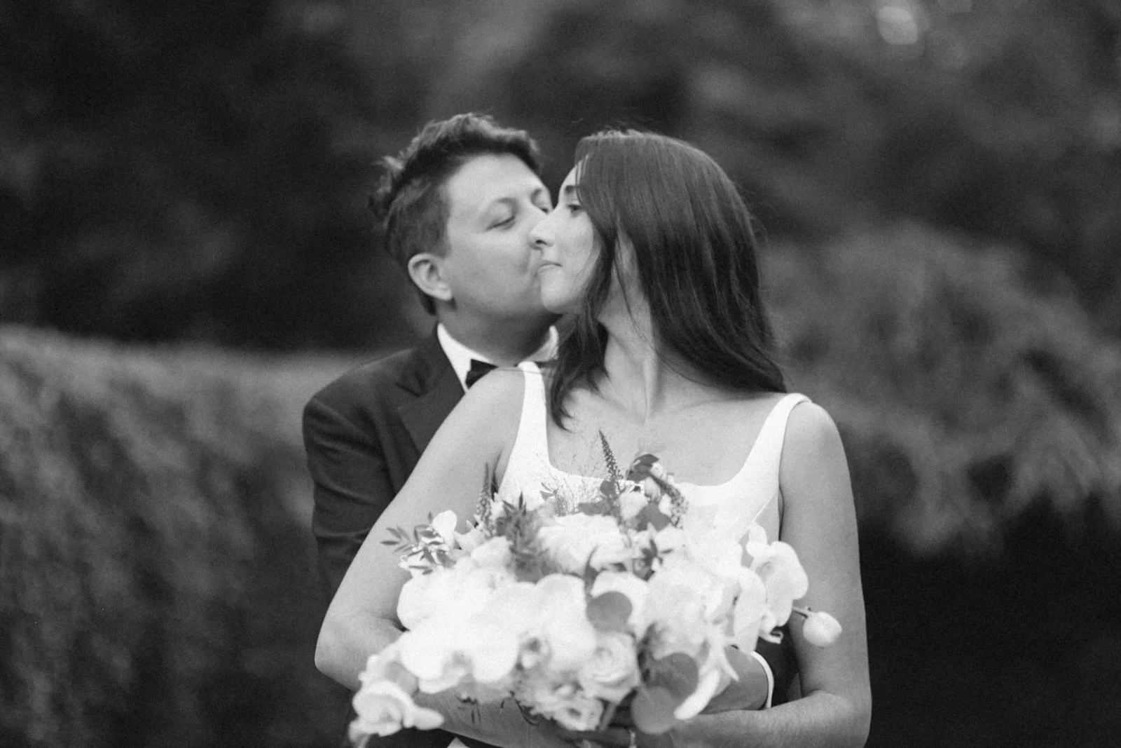 A black and white photo of a couple kissing, with the woman holding a bouquet of flowers. The man is dressed in a tuxedo, and the woman is wearing a wedding dress.