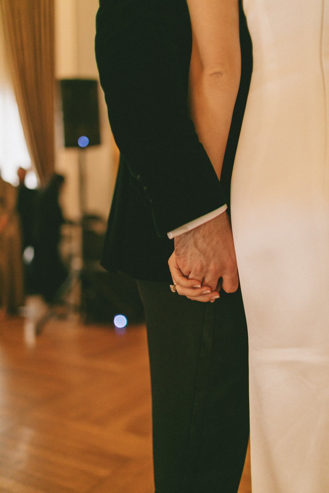 Close-up of two people holding hands, one wearing a black suit and the other wearing a white shirt and black pants, in a warmly lit room.