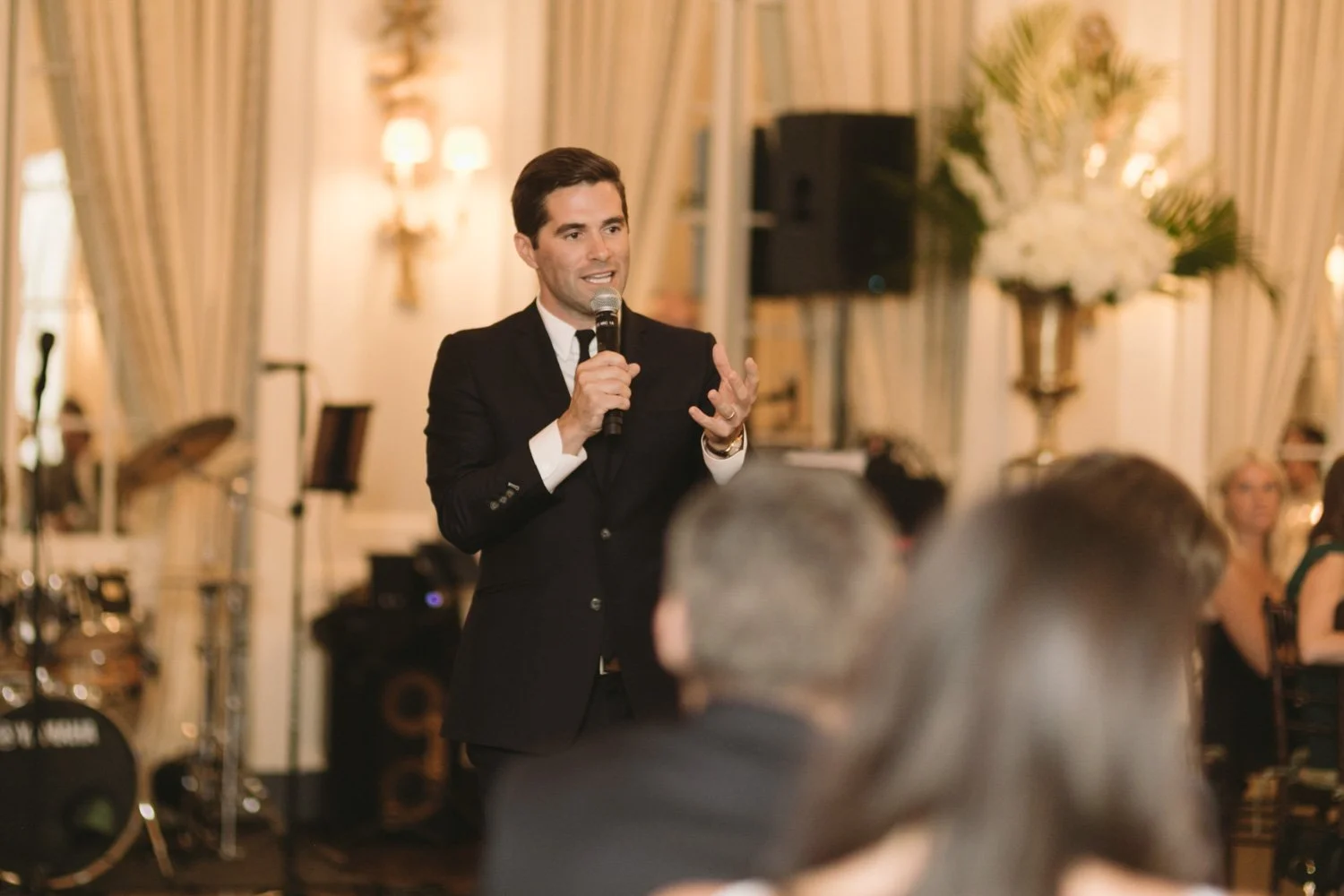 A man in a black suit giving a speech with a microphone in a decorated event hall.