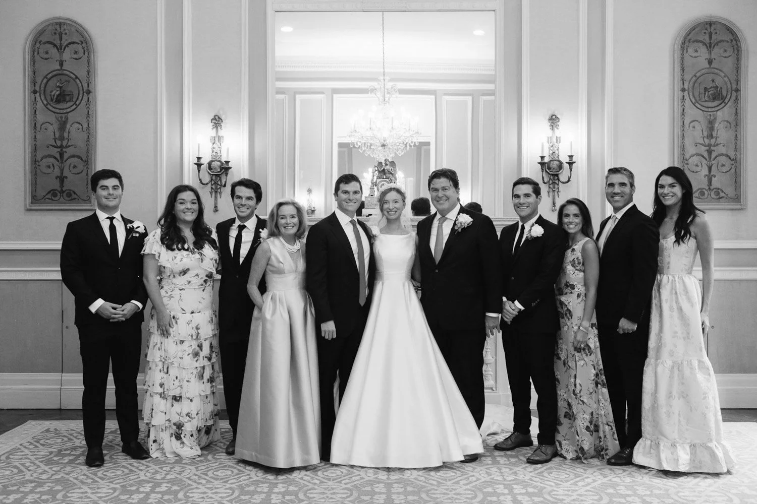 Group of people at a wedding, with bride in a white gown and groom in a dark suit in the center, surrounded by friends and family, inside a decorated elegant room.