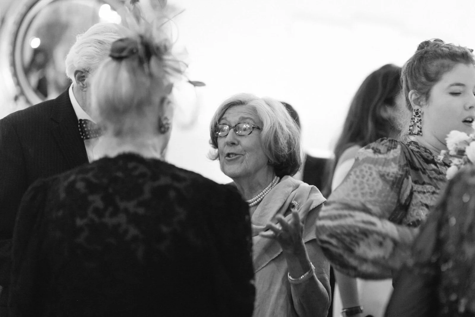 A group of people, mostly women, socializing in elegant attire at what appears to be a social or formal event. One elderly woman with glasses is speaking animatedly to another woman with styled hair and patterned clothing.